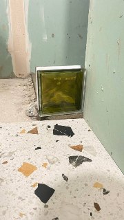 Фото угла ремонта: стеклоблок внизу, зелёный гипсокартон на стенах, ободранный пол с терраццо-плиткой и строительная пыль