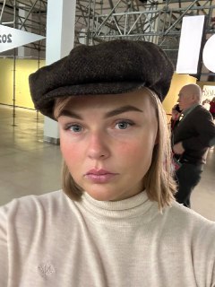 Портретная фотография молодой женщины в кепи и светлом свитере на фоне выставочного зала с потолочными фермами, стендами и посетителями