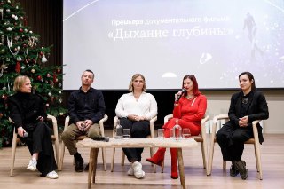 Панель с несколькими людьми, сидящими на сцене перед большим экраном с названием фильма «Дыхание глубины» во время закрытой премьеры, видна праздничная декорация.