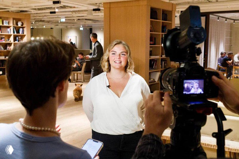 Ольга Маркина даёт интервью перед камерой на премьере: портрет в белой блузке, оператор и журналисты фиксируют репортаж в фойе мероприятия.