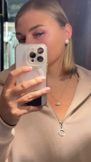 Зеркальное селфи крупным планом: человек держит телефон, видно часть лица, серьги, подвеска в бежевом топе.
