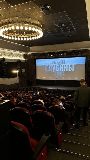 Интерьер кинозала на премьере: ряды кресел с сидящими зрителями, затемнённый зал и экран с заголовком фильма.