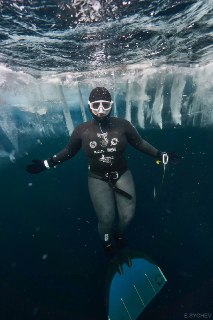 Фридайвер в гидрокостюме и маске плывет подо льдом, видна ледяная поверхность сверху, холодная подледная сцена.