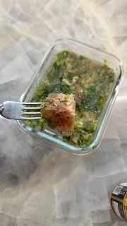 Стеклянная форма с холодцом на столе: кусок мясного студня на вилке над зелёным бульоном с мелкой зеленью и специями.