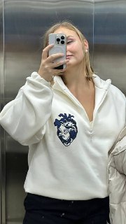Автопортрет в зеркале: женщина в белой толстовке с тёмным принтом на груди, держит телефон, виден интерьер лифта.