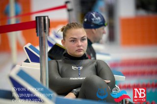 Спортсменка в гидрокостюме сидит у бортика бассейна, сосредоточенная; на заднем плане другой пловец и элементы соревнований