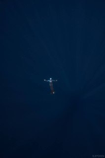 Вид сверху на одного фридайвера в глубокой тёмно-синей воде; человек небольшой на фоне масштаба моря, подчёркнуты лучи света и глубина.