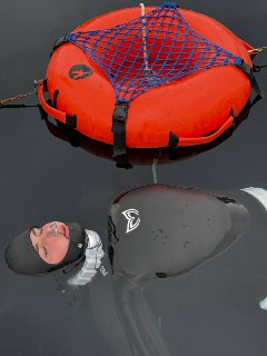 Фридайвер в чёрном гидрокостюме с капюшоном лежит у поверхности воды рядом с ярко‑оранжевым спасательным буйком и сеткой.