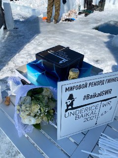 Зимняя сцена у проруби: на столике лежат коробка, букет и табличка о мировом рекорде Гиннесса, вокруг снег и видны люди.