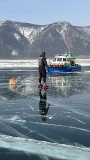 Человек на льду рядом с Бухтой, вдалеке видно судно на поверхности Байкала и гористый берег, отчётливые трещины на льду.