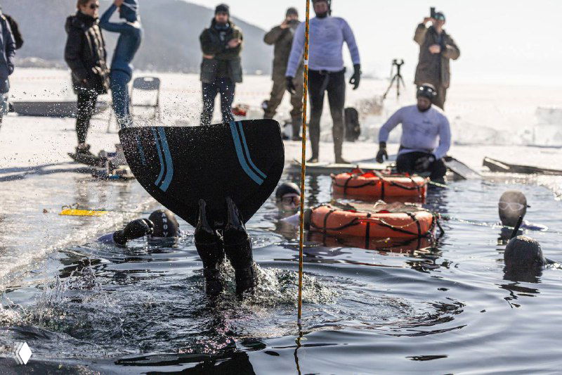 Фридайвер выходит через прорубь на Байкале, команда поддержки и спасательное снаряжение стоят на льду рядом с открытой водой и плотом.