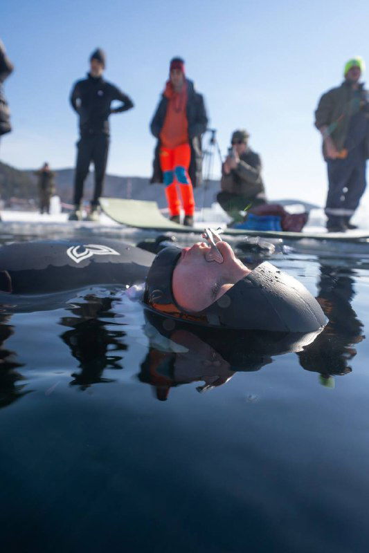 Фридайвер расслабленно лежит на поверхности воды в гидрокостюме у проруби, на льду позади видны наблюдающие и поддержка соревнований.