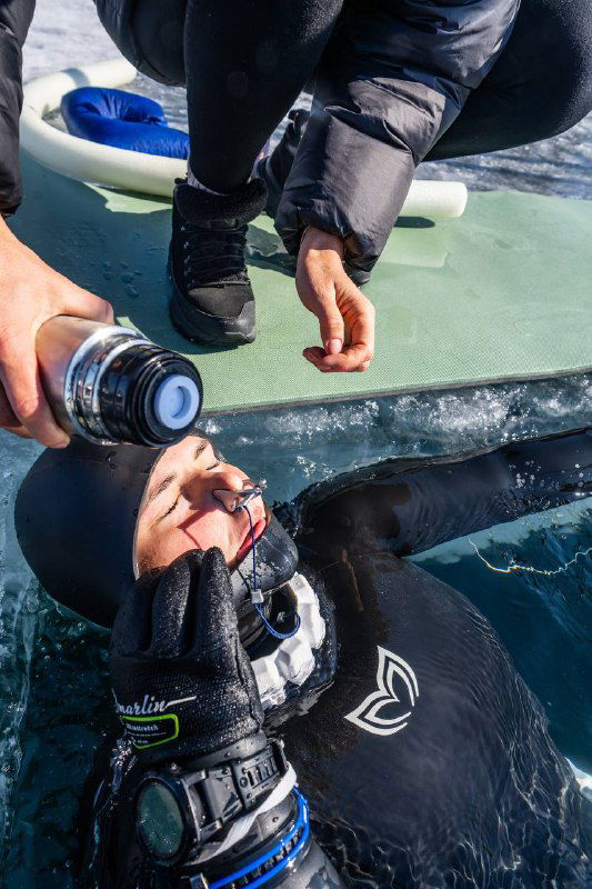Инструктор подаёт дыхательное устройство фридайверу на плоту рядом с прорубью, руки, регулятор и поддерживающее оборудование в кадре.