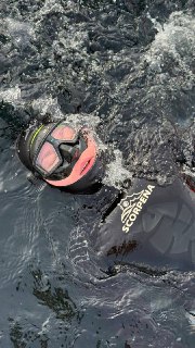 Фридайвер в гидрокостюме с маской частично погружён в холодную воду Байкала, видны защитные приборы и брызги рядом.