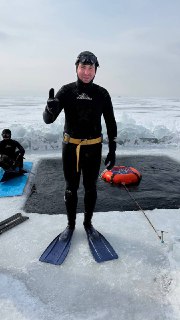 Фридайвер в гидрокостюме стоит на льду в ластах и снаряжении, показывает жест одобрения, фон — ледяные просторы Байкала.