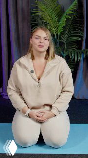 Ольга Маркина сидит на коленях на коврике в студии и объясняет технику диафрагмального дыхания; на фоне видна пальма и шторы.