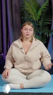 Ольга Маркина сидит на коврике в студии и показывает технику диафрагмального дыхания; позади — шторы и зеленое растение.