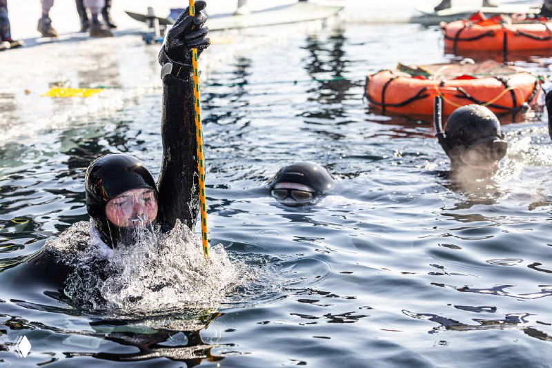 Фридайверы в воде перед нырком: один ныряльщик поднимает руку с ланьярдом, видны спасательные буи и другие участники тренировки в гидрокостюмах.