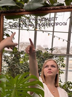 Женщина в теплице у зеркала, указывает рукой вверх; вокруг листья растений, отражение в стекле и конструкция теплицы.