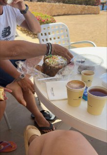 Стол на веранде с пасхальным куличом в упаковке, несколькими бумажными стаканчиками и частично видимыми руками людей.