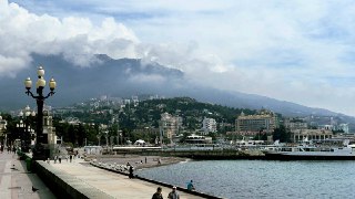 Вид на прибрежную набережную Крыма: спокойное море, прогулочная зона с фонарями, здания у подножия гор и облачное небо.