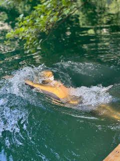 Плавец в озерной воде: виден сильный всплеск и движение, зелёная поверхность воды и размытый прибрежный растительный фон вокруг.