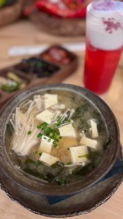 Глубокая миска мисо‑супа с кубиками тофу, пучками эноки и зелёными листьями, на фоне видно высокий розово‑белый напиток и поднос с закусками.