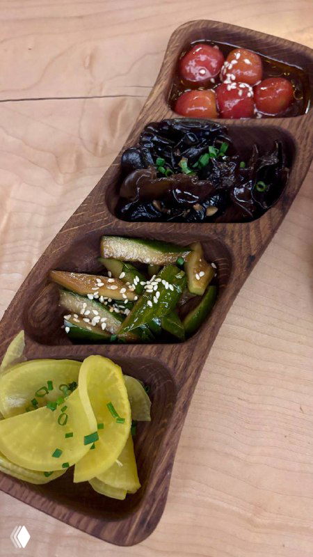 Деревянная разделённая тарелка с четырьмя типами цукэмоно: маринованный дайкон, огурцы, грибы и томаты, украшенная кунжутом и зеленью.