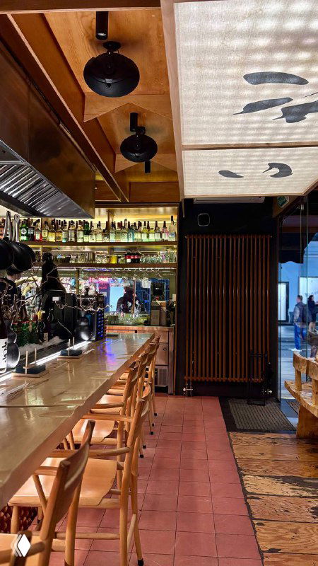 Интерьер ресторана: длинная барная стойка, высокие стулья и японская вывеска — атмосферное вечернее пространство заведения.