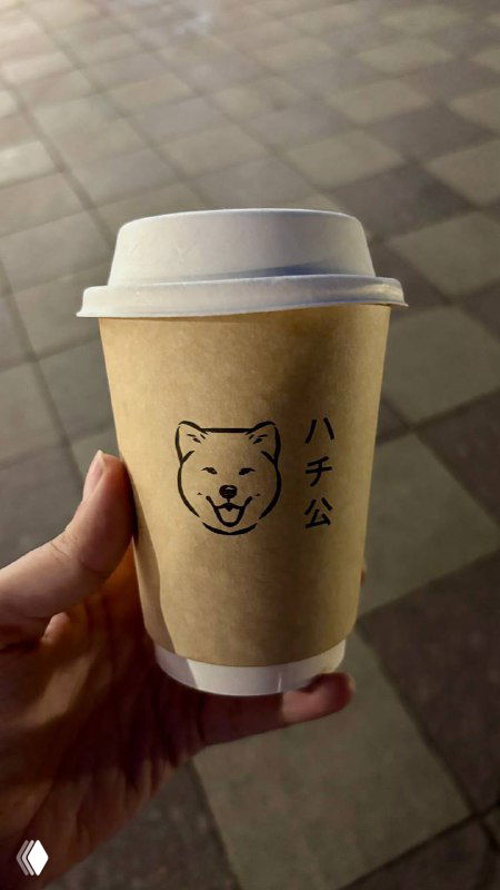 Крафтовый бумажный стакан с логотипом Хатико в руке на фоне городской улицы — напиток навынос вечером возле ресторана.