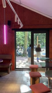 Интерьер заведения с большими стеклянными дверями, розовой неоновой лампой и видом на террасу, посетители у входа и мягкое внутреннее освещение.