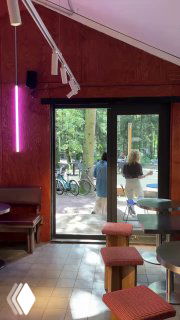 Интерьер кафе с видом на выход в лес: барные стулья, большие двери и прохожие у велосипедов на летней площадке за стеклом.