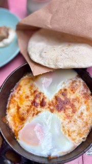 Сковородка с запечёнными яйцами и расплавленным сыром, рядом видна пита и детали сервировки — кадр с блюдом шакшука в кафе