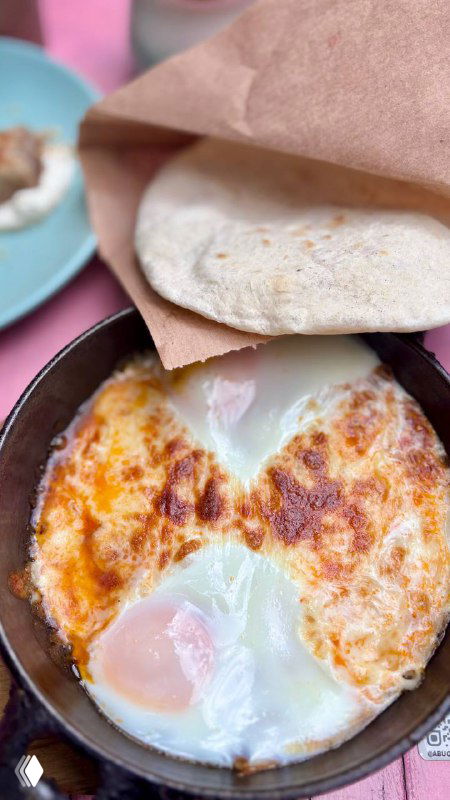 Чугунная сковорода с шакшукой: два запечённых яйца в расплавленном сыре, рядом сложена пита и виден розовый стол — интерьер кафе.
