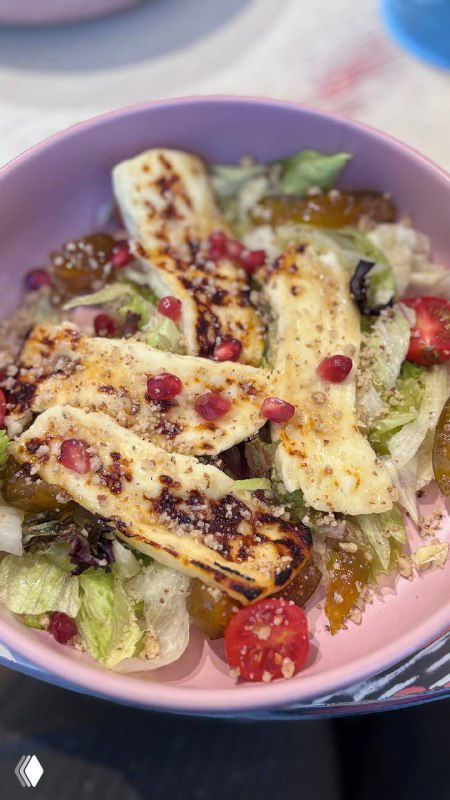 Салат с жареным халуми: полоски сыра на листах салата, сверху зерна граната и черри, в розовой тарелке на цветном столе.