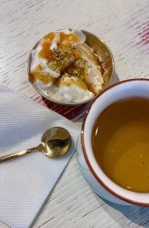 Стакан с чаём и небольшой кремовый десерт в чашке, рядом ложка и салфетка — кадр молочного пудинга с фисташками и чаем