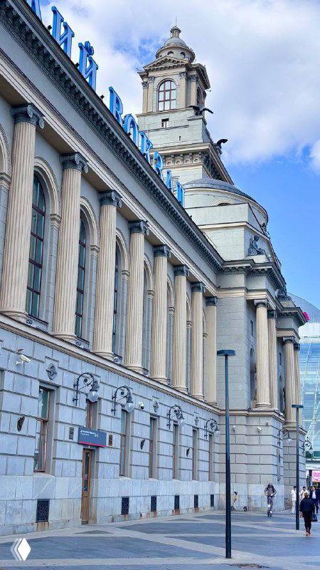 Фасад Киевского вокзала с колоннами и высокой часовой башней на фоне голубого неба; городской тротуар и прохожие на переднем плане в Москве.