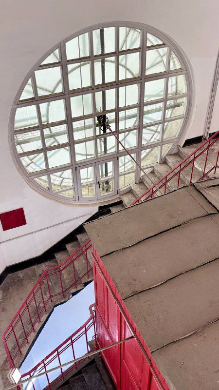 Овальное световое окно и металлическая лестница в шахте башни: вид вниз по лестнице, красные перила и конструкция смотровой площадки у часов.