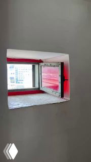 Небольшое квадратное окно с красной рамой в комнате башни, вид на внутреннее пространство и фрагмент стены с покраской и вентиляционным проемом.