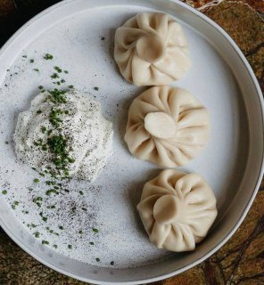 Тарелка с тройкой хинкали и порцией кремообразного соуса, подача на светлой керамической тарелке на деревянном фоне.