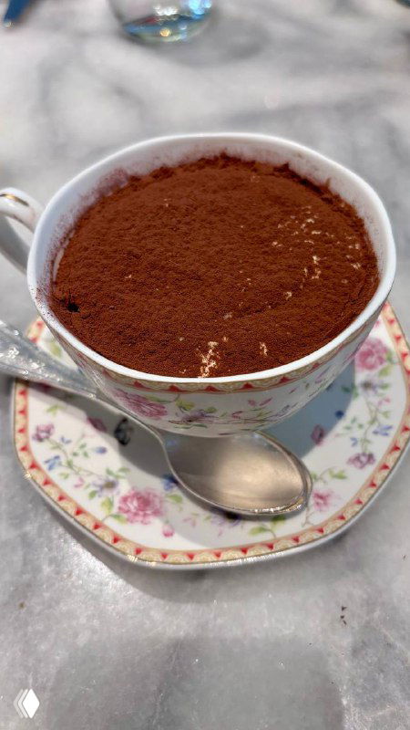 Тирамису в чашке сверху посыпано какао; десерт подан в фарфоровой чашке с блюдцем и ложкой рядом на мраморном столе.