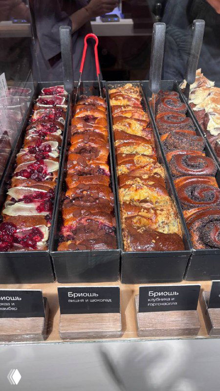 Витрина пекарни с рядами бриошей в формочках трёх вкусов; видны яркие начинки, глянцевая поверхность и наклейки с названиями.