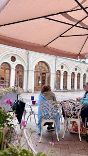 Летняя терраса буфета с гостями за белыми металлическими столиками под зонтом, арочные окна монастырского корпуса на заднем плане