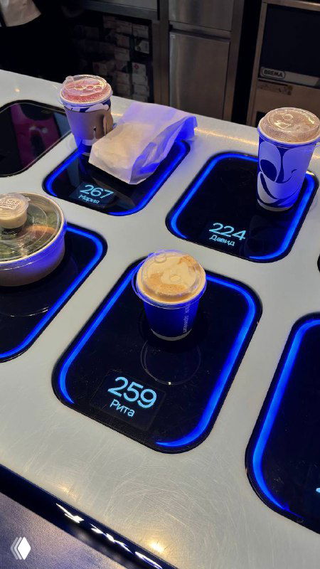 Подача напитков в кофемате: несколько бумажных стаканов с сырной пенкой стоят в подсвеченных ячейках автомата на стойке кофейни.