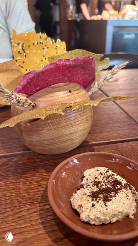 Тарелка с кремовой намазкой и хрустящей декоративной стружкой на столе бистро; часть сервировки и фоновая атмосфера заведения.