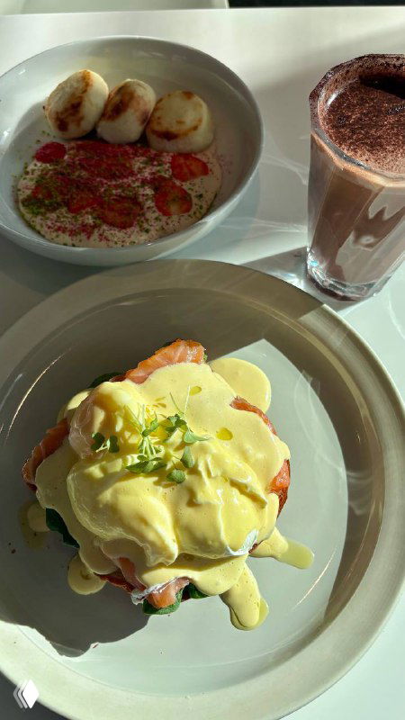 Завтрак в rockets: яйцо-пашот на бриоши на первом плане, рядом тарелка с сырниками и высокий стакан горячего напитка — интерьер кафе на заднем плане.