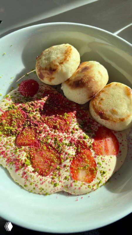 Тарелка с сырниками и ягодами: сверху воздушный крем из сгущенки, посыпка и кусочки инжира рядом, солнце падает через окно кафе.