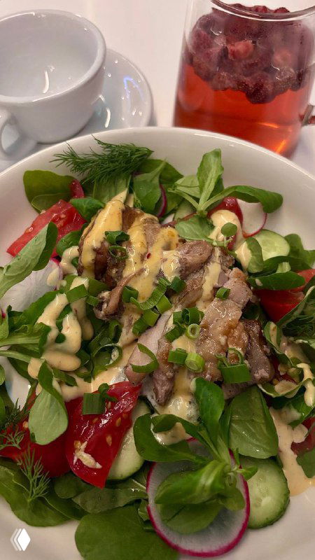 Большая тарелка салата с миксом зелени, ломтиками овощей и заправкой; на заднем плане стакан с травяным чаем цвета ягод.