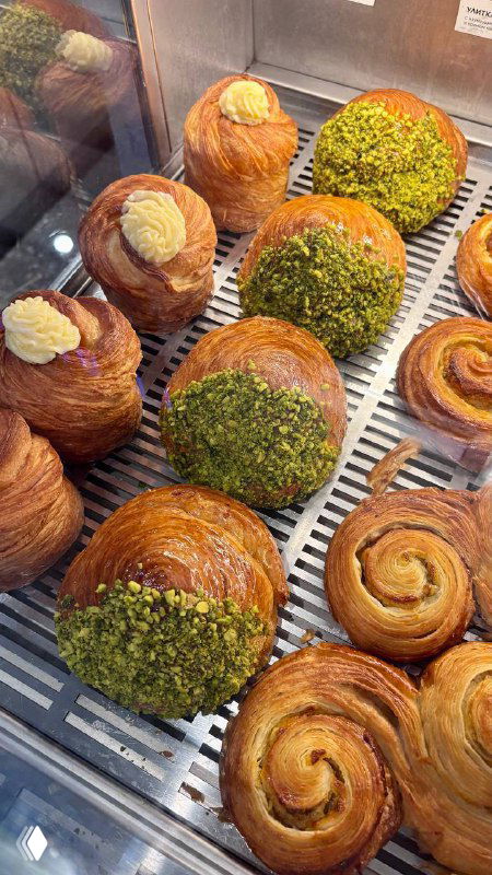 Витрина с булочками: даниши, круассаны и изделия с фисташковой посыпкой и кремовыми начинками — выпечка выложена на решётке кафе.