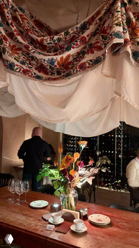 Общий вид стола с цветочной композицией, драпировками и гостями на заднем плане — настроение уютного вечера в ресторане.